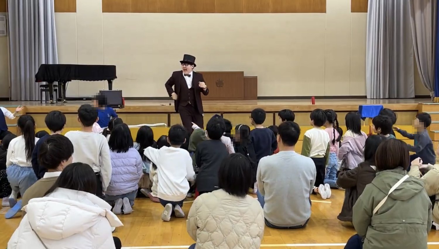 体育館の舞台上でマジックを演じるマジシャンDaiSukeと、床に座って見ている小学生と保護者の皆様