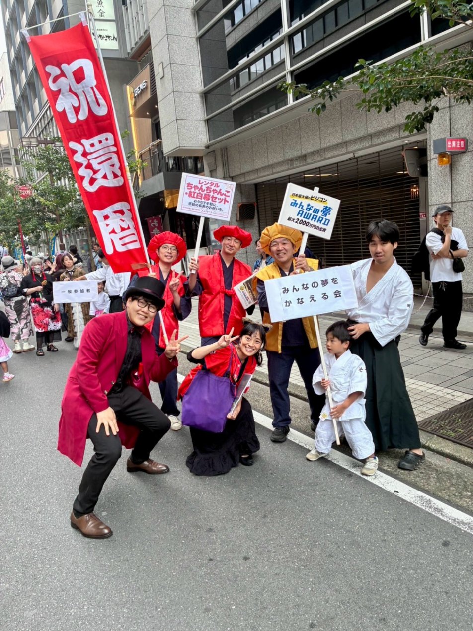 「祝還暦」ののぼりの前で、赤いジャケットにシルクハット姿のDaiSukeがポーズを取り、一緒に練り歩く仲間たちと並ぶ集合写真