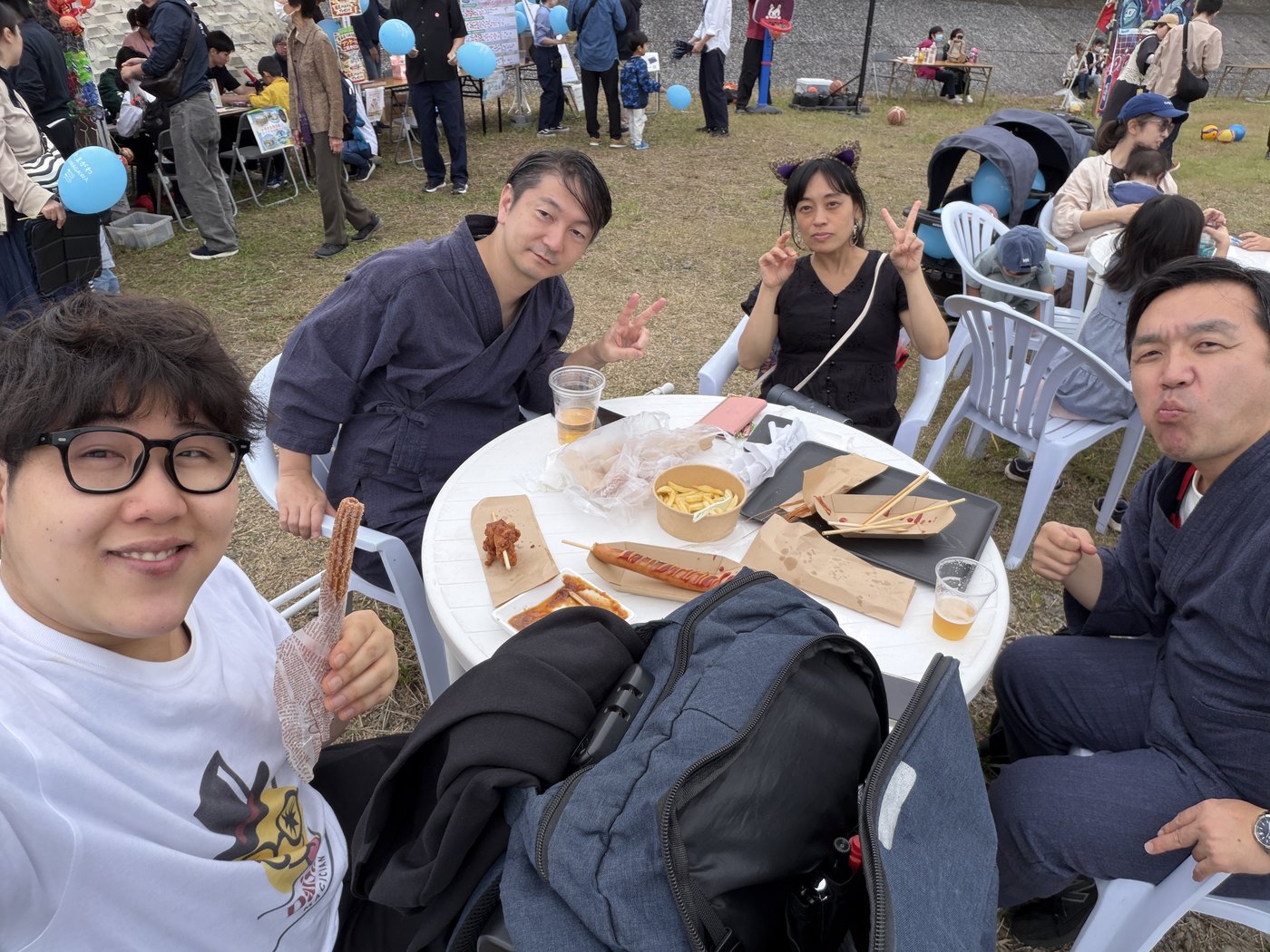 野外会場のテーブルで、屋台の料理を囲んで仲間たちとくつろぐ私服姿のDaiSuke
