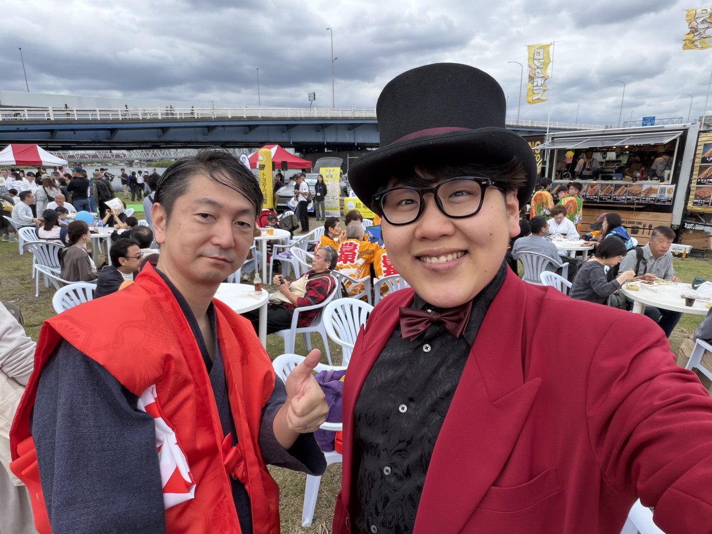 屋台が並ぶ野外会場で、赤いジャケットにシルクハット姿のDaiSukeが仲間と並んだ自撮り