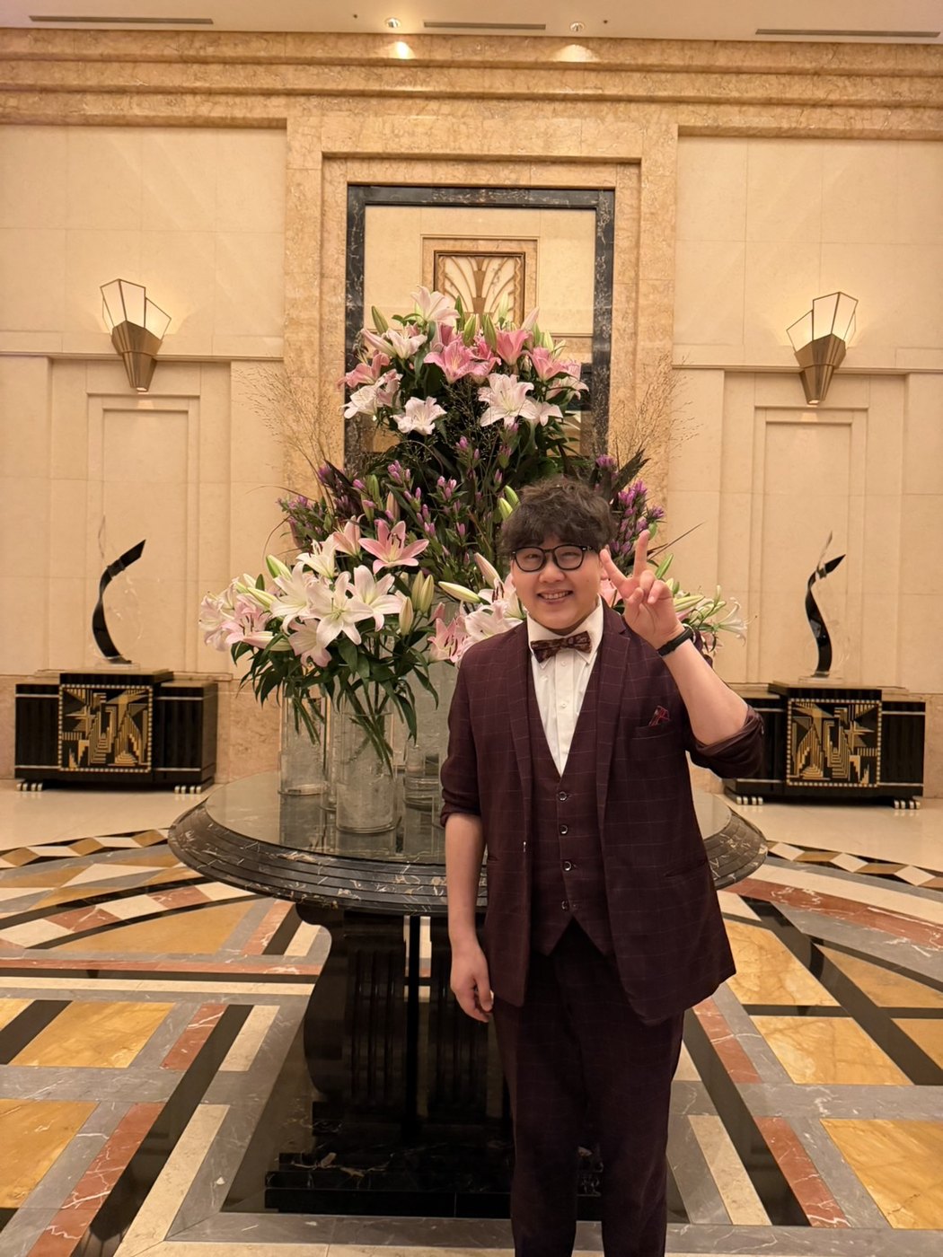 大理石の床と大きな花のアレンジメントが飾られたホテル館内で、ピースをするDaiSuke
