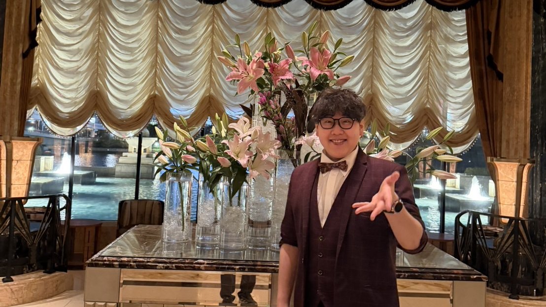 ドレープカーテンとピンクのユリが飾られたホテルのロビーで、手を差し出してポーズをとるDaiSuke